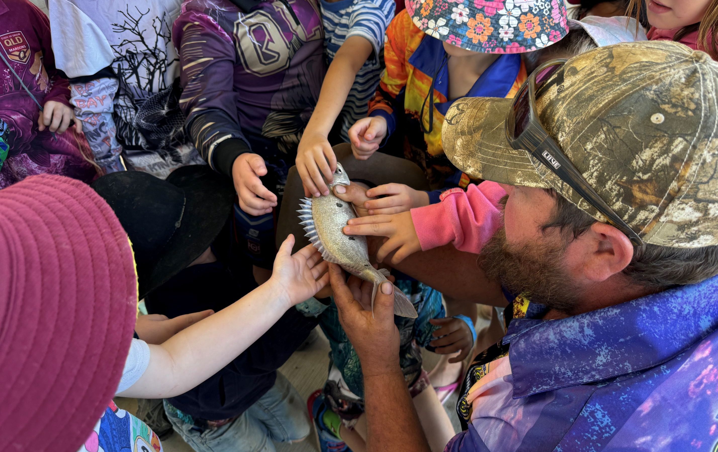 Inspecting a fish at Longreach Junior Fishing Day Children gather around, hands stretched out to touch the scales of a silver fish.
