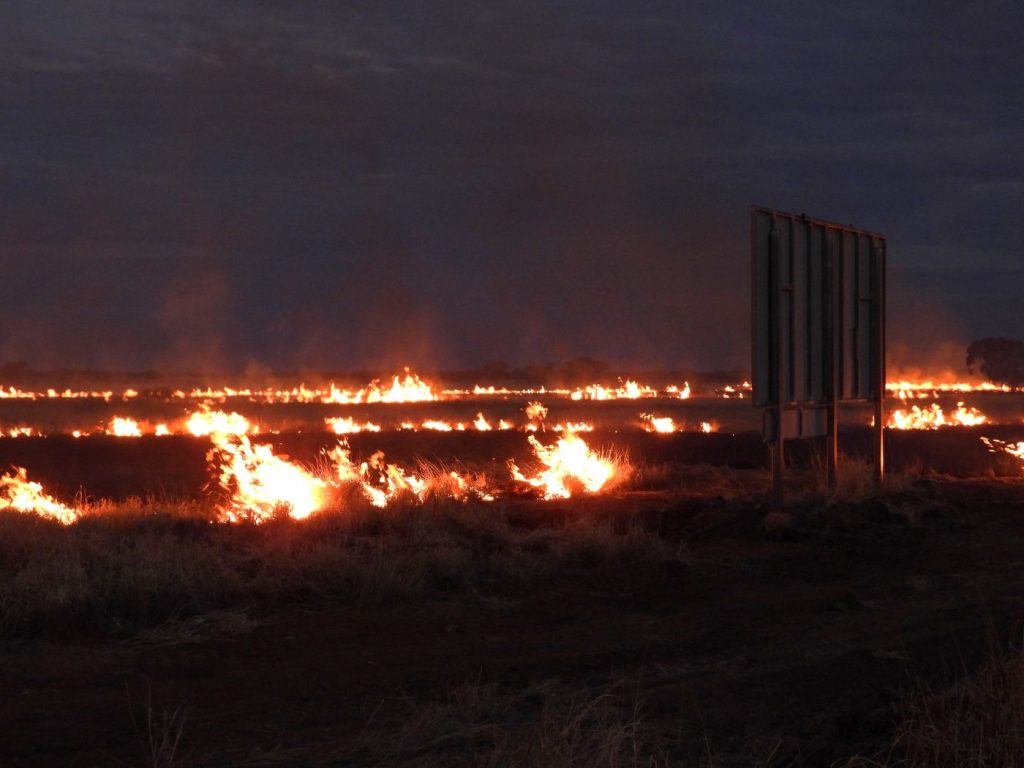 A sign is sillhouetted against the flames of a large fire on stock route at dusk.